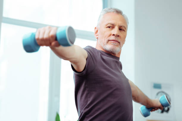Strong fit aged man looking at his hands Developing muscles. Strong fit aged man looking at his hand while training with dumbbells exercise room photos stock pictures, royalty-free photos & images