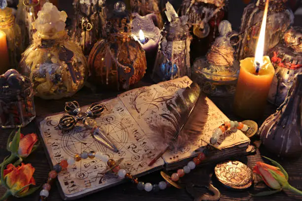 Photo of Witch book with cross, quill, burning candles and magic bottles on the table.