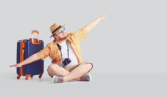 Fat funny man with a beard in a summer shirt in a hat with a suitcase raised his hands up the airplane symbol on a gray background. The original concept of recreation, travel in the summer.