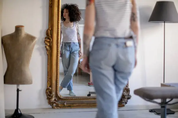 Reflection of a young tattooed woman with brown curly hair standing in front of the mirror and looking away