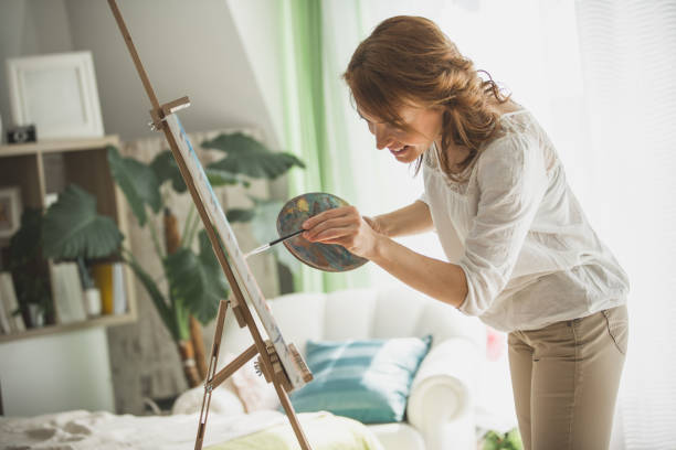 Happy mid adult woman enjoying while doing her favorite hobbie, painting. She is releasing her creativity on canvas and smiling.