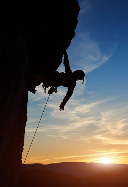 istock Male climber silhouette hanging on rock, one hand down. Risky activity in mountains at sunset on cloudy sky. Copy space 1148998556