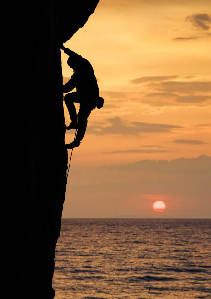 istock Silhouette of man rock climbing on cliff at sunset above ocean. Side view. Sun on orange sky on background. Copy space 1148998564