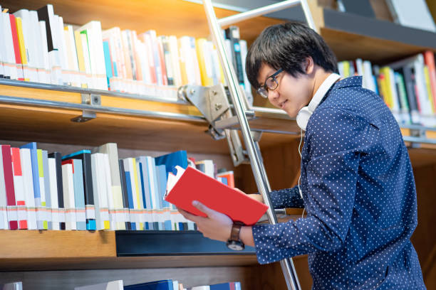 istock Asian man university student choosing book from bookshelf while standing on the ladder in library for education research. Bestseller collection in bookstore. Scholarship or educational opportunity 1149011084