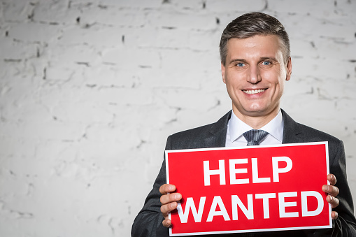 Portrait of smiling businessman holding help wanted sign while standing against white brick wall