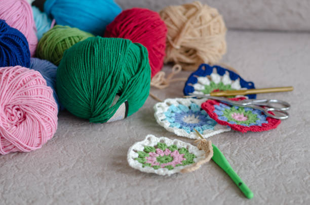 Multi Colored plaid squares of crochet on a cream colored seat background. Green, pink,cream, blue,red color yarns,hookeds, scissors are  on the sofa.