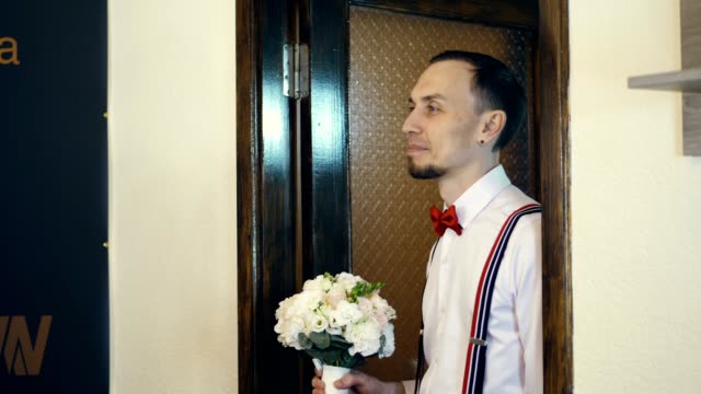 in the doorway the young guy comes in, the groom, in a red bow tie and with a bouquet of flowers. he looks at the bride