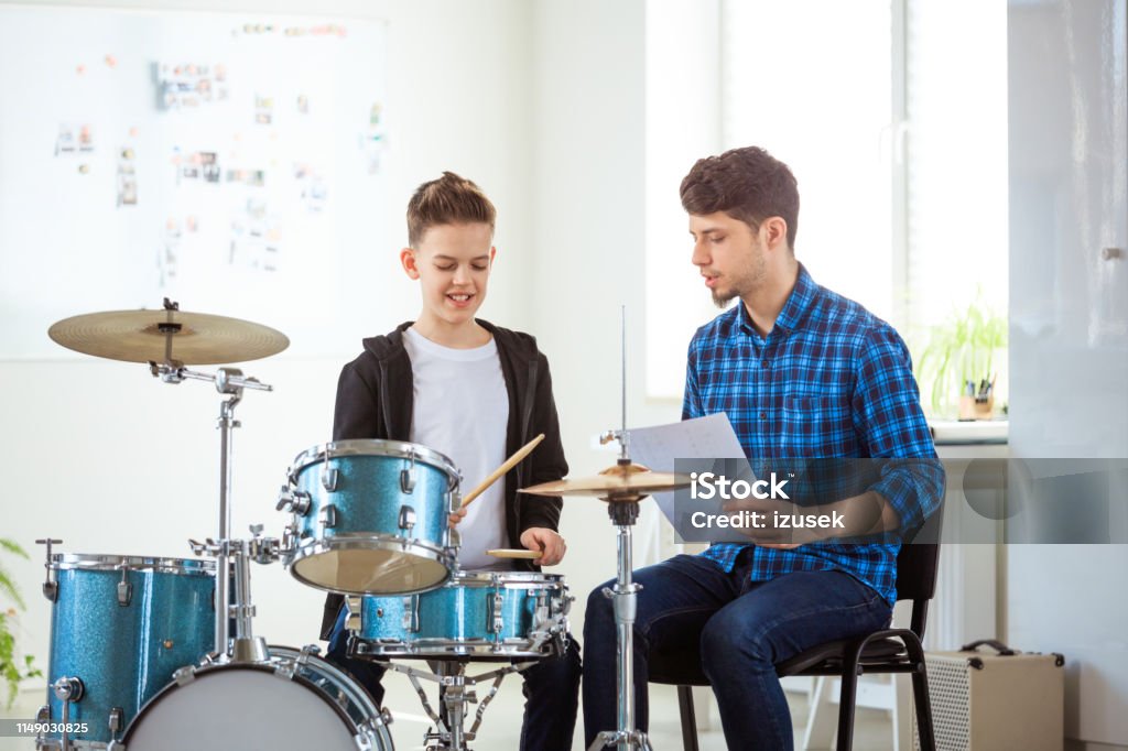 Teacher showing sheet to boy while playing drums Mid adult teacher showing music sheet to pre-adolescent boy. Little drummer is practicing with male instructor. They are sitting in conservatory. 12-13 Years Stock Photo