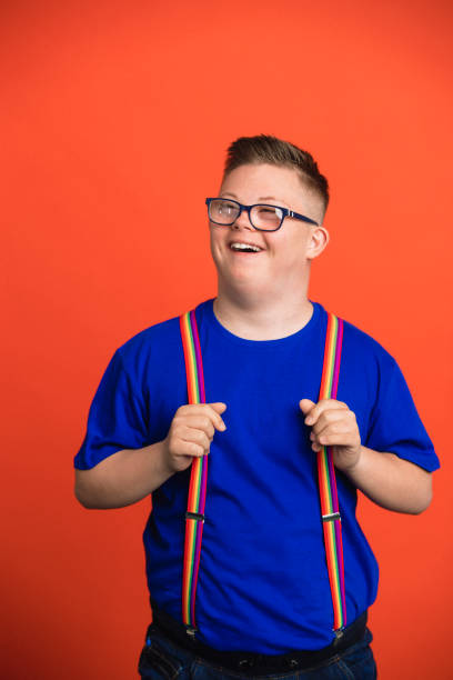 Fun in the Studio Teenage boy with Down Syndrome standing in front of an orange studio background laughing as he looks away from the camera, pulling his suspenders away from him. down syndrome photos stock pictures, royalty-free photos & images