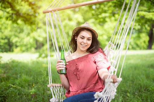 Young beautiful Latino woman relaxing on hammock outside, holds a glass beer bottle and smiling.