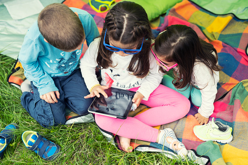 Three children with colorful sunglasses playing with a digital tablet on a picnic blanket under a camping tent.