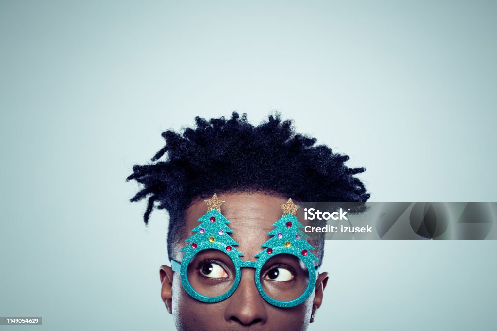 African man with christmas mask Close up of african man wearing christmas tree shaped glasses looking away on gray background. Christmas Stock Photo