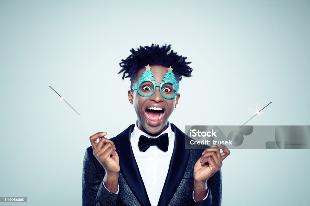 Funny christmas portrait of man with sparklers Funny christmas portrait of excited young man with sparklers. African man wearing christmas tree shaped glasses holding sparklers in both hands on gray background. Men Stock Photo
