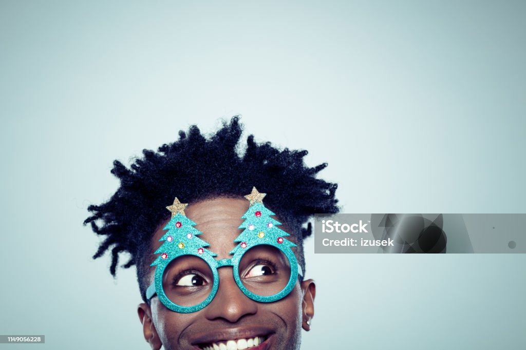 Funny christmas portrait of young guy Funny christmas portrait of young guy on gray background. Half face of afro american man wearing christmas tree shaped glasses and looking at copy space. Christmas Stock Photo
