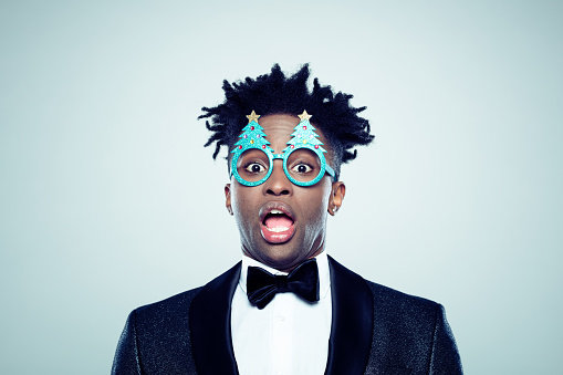 Close up portrait of african man wearing christmas tree glasses looking shocked on gray background.