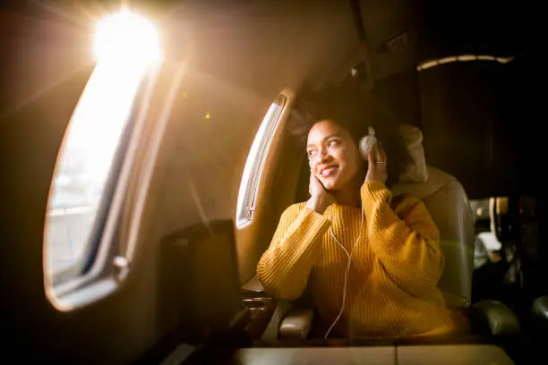 Photo of Young modern woman sitting in a private jet, listening to music through the headphones and looking through the window