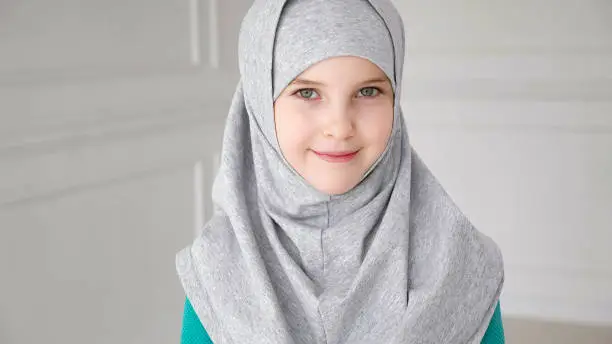 Photo of Muslim girl in grey hijab is looking in camera and laughing.