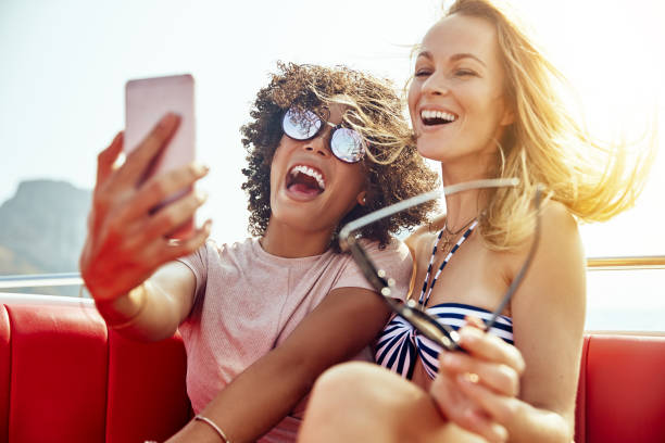 Two laughing young women sitting together on a boat taking selfies during summer vacation