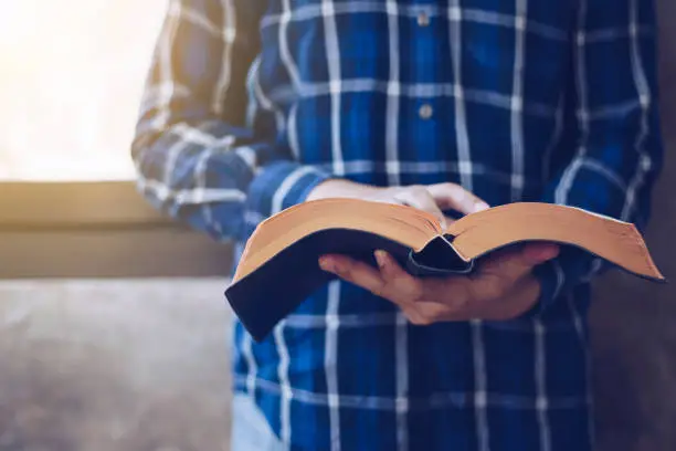 Close up of Young christian man standing at concrete wall while read bible with window light