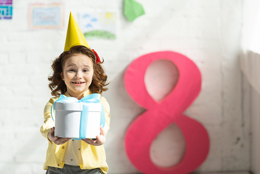adorable smiling kid in party cap holding present and posing near decorative pink number 8