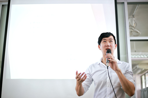 Portrait of young handsome Asian male speaker publicly speaking on stage to group of audience with white board behind.