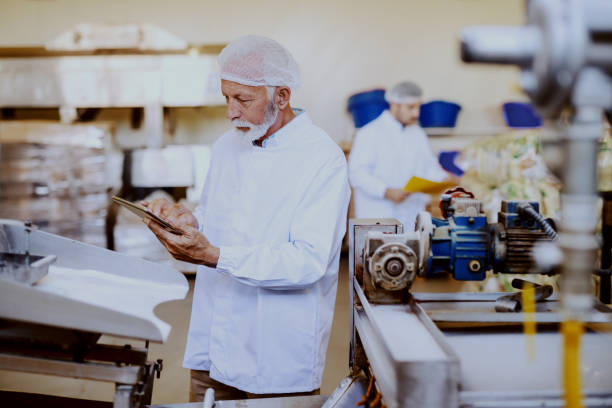 istock Senior adult supervisor in white sterile uniform checking on machine and using tablet. Food plant interior. 1149077933