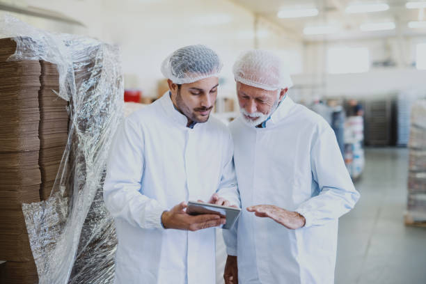 istock Young serious manager in sterile uniform using tablet for checking goods and talking with employee. Food factory interior. 1149078017