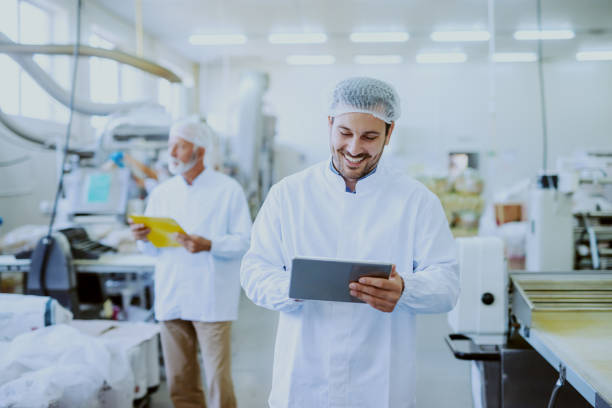 giovane supervisore sorridente caucasico in uniforme bianca sterile usando compressa mentre si trova in una pianta alimentare. in background vecchio un lavoratore che controlla la macchina. - food processing plant manufacturing factory food foto e immagini stock