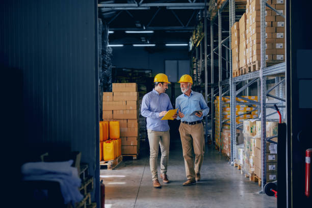 dos socios comerciales en ropa formal y con cascos amarillos protectores en cabezas caminando y hablando de negocios. más joven una carpeta de retención con datos mientras que uno más viejo usando la tableta. - manufacturar fotografías e imágenes de stock