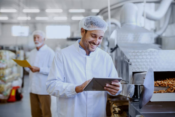 istock Young smiling supervisor in sterile white uniform using tablet and checking quality of salty sticks. In background older supervisor holding folder with documents. Food plant interior. 1149078055