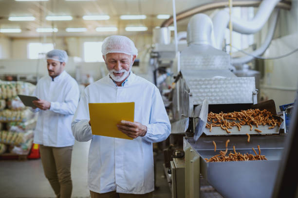 istock Caucasian senior adult quality controller holding folder with documents and checking on quality of salty sticks. Food plant interior. 1149078105