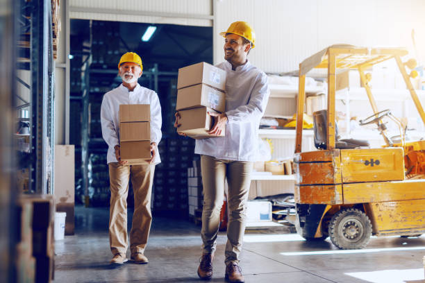 istock Two blue collar workers in white uniforms and with yellow helmets on heads relocating heavy boxes in warehouse. 1149078639