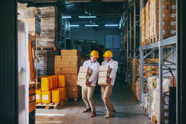 istock Two blue collar workers in white uniforms and with yellow helmets on heads relocating heavy boxes in warehouse. 1149078689