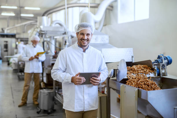 joven sonriente gerente de uniforme estéril sosteniendo la tableta y mirando a la cámara mientras está de pie en la fábrica de alimentos. - sales manager fotografías e imágenes de stock