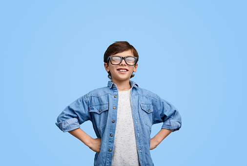 Smart content kid holding hands on waist and smiling at camera against blue backdrop