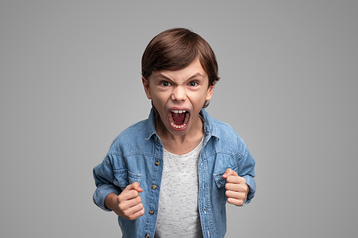 Boy in casual outfit clenching fists and shouting angrily at camera while standing on gray background