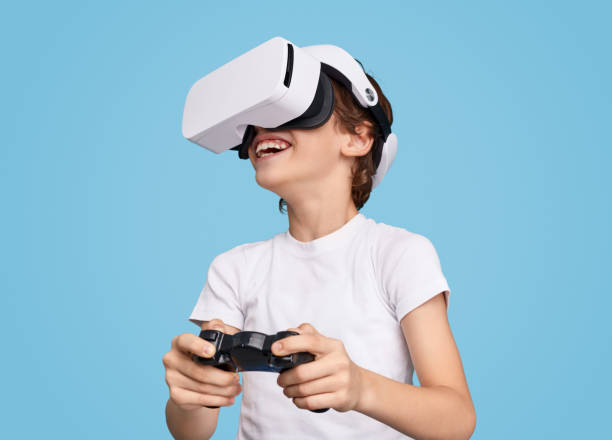 Cheerful boy playing videogame in VR goggles Happy boy enjoying virtual game while wearing VR headset and using gamepad on blue backdrop virtual reality gaming stock pictures, royalty-free photos & images