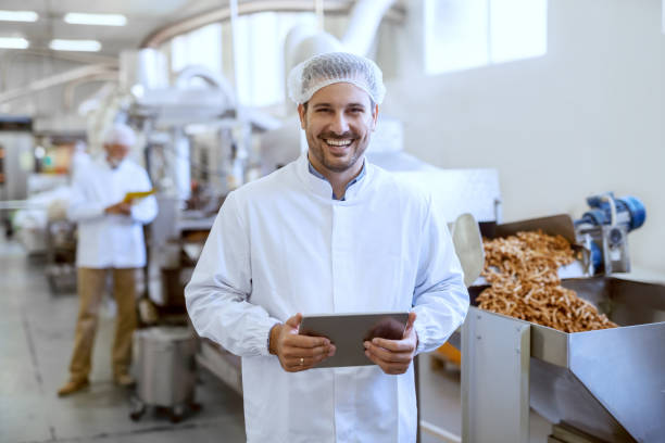 無菌の制服の若い笑顔マネージャーは、タブレットを保持し、食品工場に立っている間にカメラを見ています。 - food processing plant manufacturing factory food ストックフォトと画像