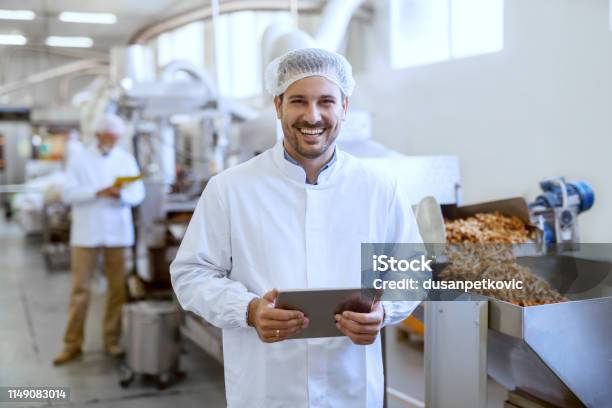 Young Smiling Manager In Sterile Uniform Holding Tablet And Looking At Camera While Standing In Food Factory Stock Photo - Download Image Now