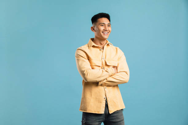 Confident and Carefree A young man standing alone in a studio with a blue backdrop. His arms are crossed and he's looking off to the side. sideways glance stock pictures, royalty-free photos & images
