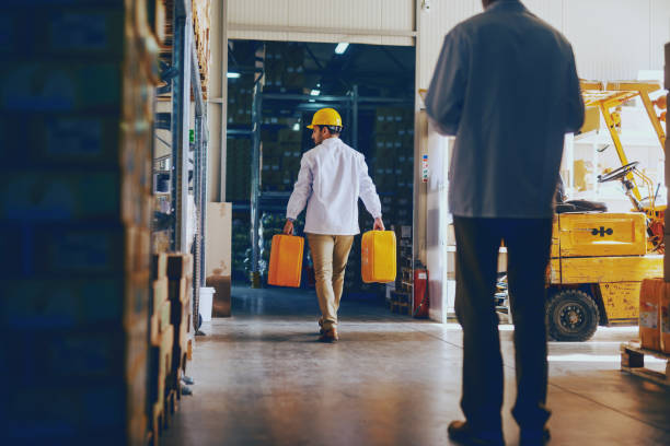 istock Young Caucasian employee dressed in white uniform and with protective helmet on head relocating oil canisters in warehouse while manager controlling him. 1149085042