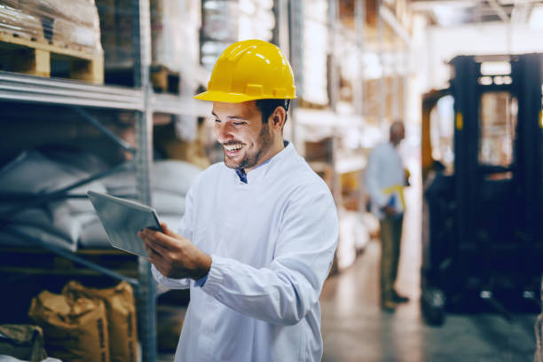 istock Young smiling Caucasian auditor in white uniform and with yellow helmet on head using tablet for checking on goods in warehouse. In background worker standing next to forklift. 1149085102