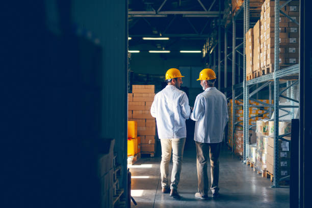 istock Two smiling warehouse workers in white uniforms and yellow helmets on heads standing and talking about job. Backs turned. 1149085173