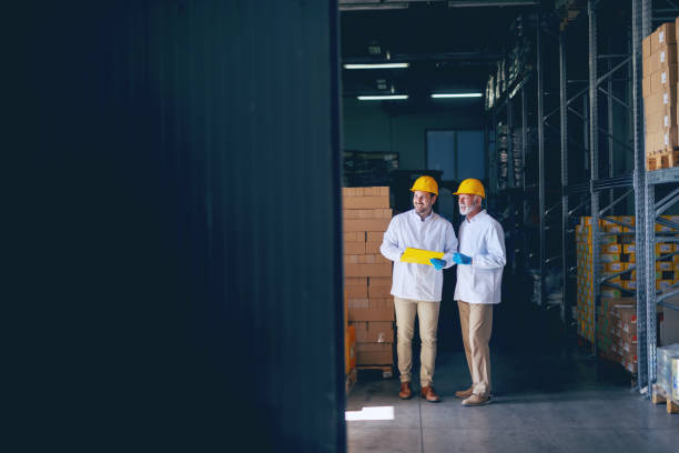 istock Two smiling warehouse workers in white uniforms and yellow helmets on heads standing and talking about job. Younger one holding folder with documents in hands. 1149085225