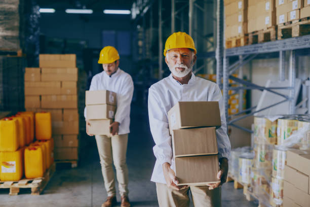 istock Two blue collar workers in white uniforms and with yellow helmets on heads relocating heavy boxes in warehouse. Selective focus on senior adult employee. 1149085426