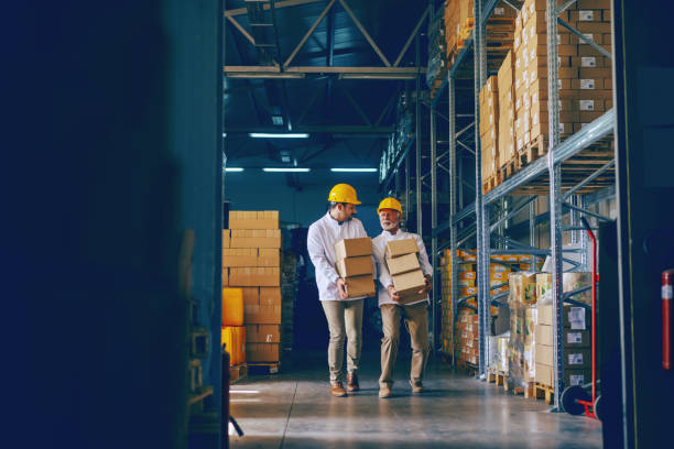 istock Two blue collar workers in white uniforms and with yellow helmets on heads relocating heavy boxes in warehouse. 1149086358