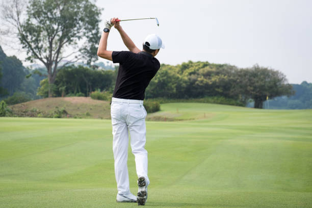 Rear view of man teeing off on golf course. Full length of male golfer is swinging a golf club at green field. He is wearing sports clothing.