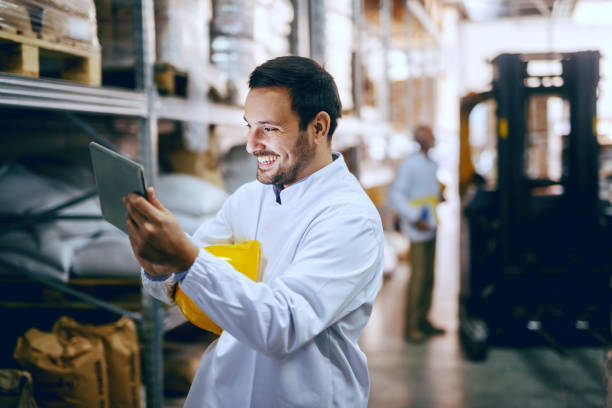 istock Smiling young Caucasian blue collar worker in white uniform and with helmet under armpit using tablet for controling delivery. In background worker standing next to forklift. Warehouse interior. 1149086667