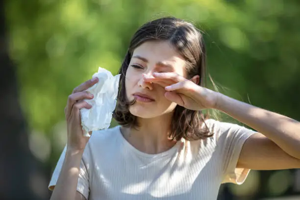 Woman Blowing Her Nose With Handkerchief In Public Parkf. Sick Young Woman With Seasonal Influenza Blowing Her Nose On A Tissue. Seasonal Virus Infection. Chronic Disease Control, Allergy Induced Asthma Remedy And Chronic Pulmonary Disease Concept.