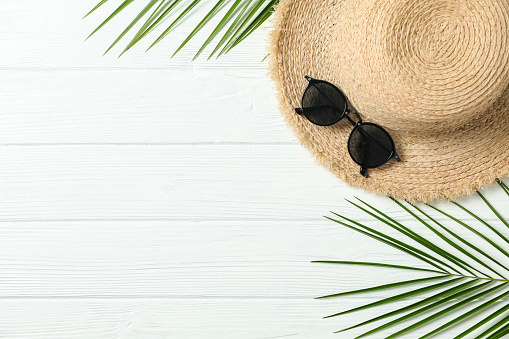 Straw hat, sunglasses and palm leaves on white wooden background, space for text and top view. Summer vacation backdrop